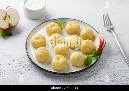 Glutenfreie gedämpfte, faule Teigtaschen mit Apfelfüllung auf einem weißen Teller, dekoriert mit Minze, umgeben von einem halben Apfel und saurer Sahne auf einem Stockfoto