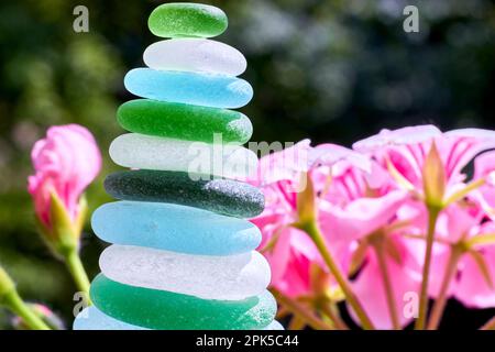 Zen-Pyramide aus Glassteinen vom Strand auf einem Hintergrund mit roten Blumen. Zen-Gleichgewicht im Garten Stockfoto