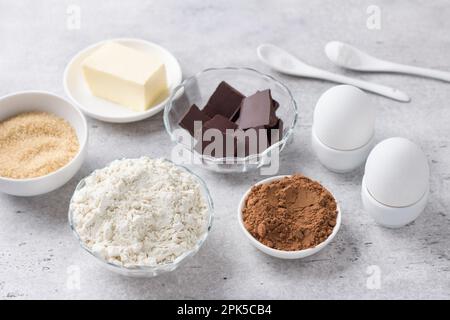 Zutaten für die Zubereitung von Schokoladenkuchen: Mehl, Schokolade, Kakao, Eier, Mandeln, Brauner Zucker, Butter auf grauem, strukturiertem Hintergrund. Köstliche Hure Stockfoto