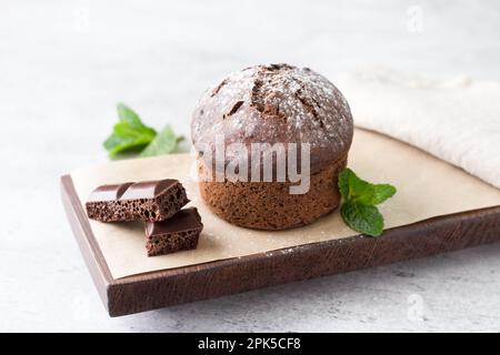 Schokoladenkuchen mit Schokoladenstückchen, garniert mit frischer Minze auf einem dunkelbraunen Brett auf grauem, strukturiertem Hintergrund. Köstliches hausgemachtes Essen Stockfoto