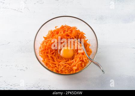 Glasschüssel mit geriebenen Karotten mit einem Ei auf einem hellgrauen Tisch, Draufsicht. Hausgemachte vegetarische Speisen: Karotten-Pfannkuchen, Pommes Frites oder andere Speisen Stockfoto