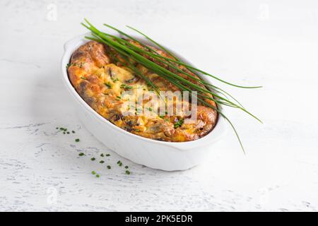 Auflauf oder Clafoutis mit Pilzen und Käse in einer weißen Keramikbackform mit grünen Zwiebeln auf hellem Hintergrund. Köstlich hausgemacht Stockfoto