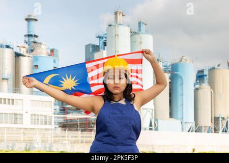 Besorgtes asiatisches Mädchen in Arbeitskleidung und Schutzhelm mit der Flagge malaysias vor der Industrielandschaft Stockfoto