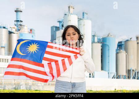 Traurige junge Frau mit der Flagge Malaysias vor dem Hintergrund der Fabrik Stockfoto