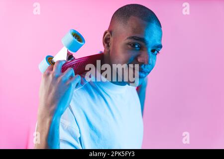Junger männlicher Skater im weißen T-Shirt hält ein kleines Skateboard am Hals und schaut auf die Kamera vor pinkfarbenem Hintergrund. Hochwertiges Foto Stockfoto