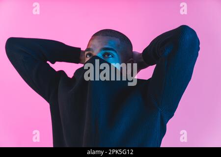 Ein cooler und modischer Mann. Der junge Schwarze versteckt sein Gesicht in seinem Sweatshirt und hält Hände hinter dem Hals. Rosafarbener Hintergrund für Studio. Hochwertiges Foto Stockfoto