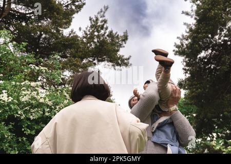 Foto einer multikulturellen Familie aus niedriger Perspektive, die mit dem Baby spielt, es hebt und das Kind in der Natur in die Luft wirft. Nicht wiedererkennbarer Vater hält Kind fest Stockfoto