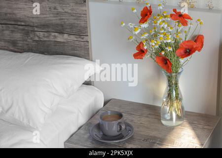 Blumenstrauß mit wunderschönen Wildblumen und Tasse mit Kaffee auf dem Nachttisch im Schlafzimmer Stockfoto