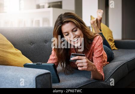 Zeit für eine Probefahrt mit dem neuen Baby. Eine attraktive junge Frau, die zu Hause ein digitales Tablet und eine Kreditkarte auf dem Sofa benutzt. Stockfoto