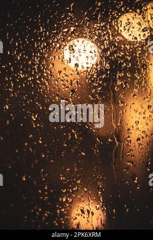 Transparente nasse Glasoberfläche mit Wassertropfen gegen helle, leuchtende Bokeh-Lichter bei dunkler Abendzeit Stockfoto