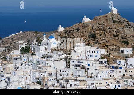 Panoramablick auf eine Kirche, kleinere Kapellen und die griechische Flagge auf einem Hügel mit Blick auf die Ägäis in iOS in Griechenland, auch bekannt als Chora Stockfoto