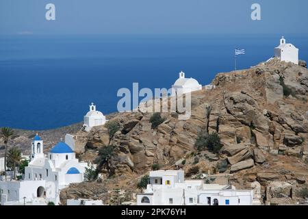 Panoramablick auf eine Kirche, kleinere Kapellen und die griechische Flagge auf einem Hügel mit Blick auf die Ägäis in iOS in Griechenland, auch bekannt als Chora Stockfoto