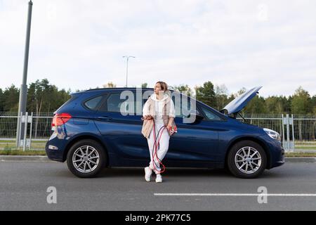 Müde Fahrerin, die ein Voltmeter auf dem Hintergrund eines kaputten Autos hält Stockfoto