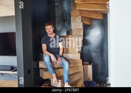 Ein Mann in lässigen Kleidern, der drinnen auf der Holztreppe sitzt Stockfoto