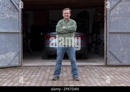 Lächelnder, erwachsener Mechaniker steht mit gekreuzten Armen vor einer Garage, aus der Nähe Stockfoto