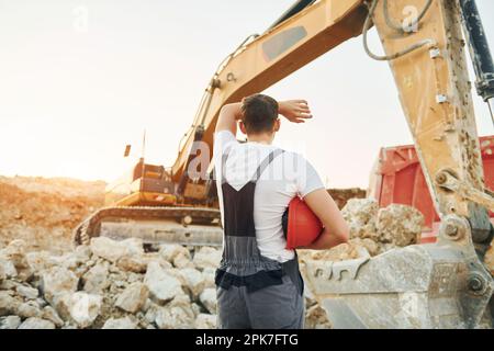 Steht gegen gelbes Fahrzeug. Arbeiter in Berufsuniform ist tagsüber auf der Leihgrube Stockfoto