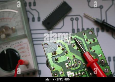 Elektronische Reparaturwerkbank. Elektronische Leiterplatte mit verschwommenem Multimeter, Chip- und Diagrammhintergrund. Auf rote Sonde fokussiert. Stockfoto