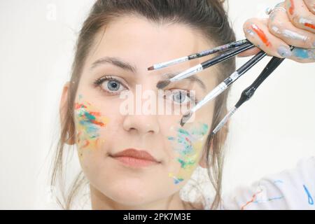 Ein Mädchen mit blauen Augen, gemalt mit bunten Farben und Bleistiften. Porträt eines Mädchens, das mit den Händen und dem Gesicht voller Farben malt. Mädchengesicht gemalt Stockfoto