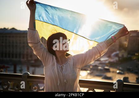 Eine bezaubernde Weiße in Silhouette in weißer Bluse und Brille hält die Flagge der Ukraine, winkt im Wind mit Blick auf die Stadt und dem Leuchteffekt der Linse. Patriotische Frau mit ukrainischer Flagge Stockfoto