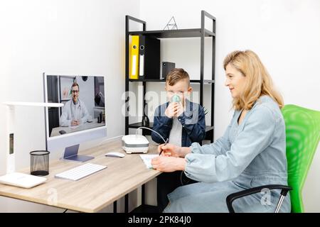 Kaukasischer kleiner Junge, der zu Hause mit einem Vernebler inhaliert. Eine junge Mutter, die ihrem Sohn beim Einatmen mit einem Vernebler hilft, bei einem Online-Meeting mit einem D. Stockfoto