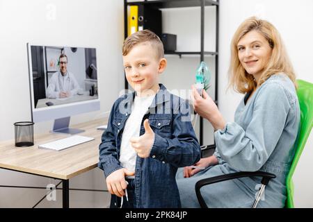 Kaukasischer kleiner Junge, der zu Hause mit einem Vernebler inhaliert. Eine junge Mutter, die ihrem Sohn beim Einatmen mit einem Vernebler hilft, bei einem Online-Meeting mit einem D. Stockfoto