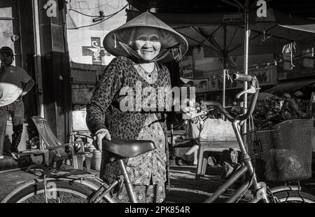 Eine ältere vietnamesische Frau, die einen konischen Hut trägt und ihr altes Fahrrad in Pleiku, Vietnam, hält. Stockfoto