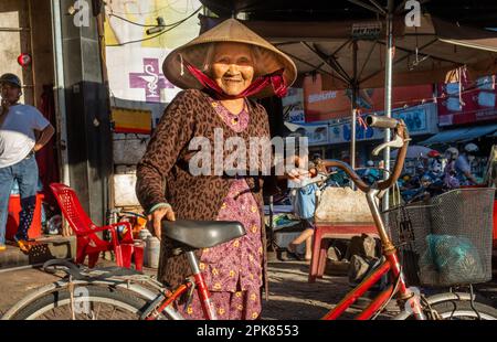 Eine ältere vietnamesische Frau, die einen konischen Hut trägt und ihr altes Fahrrad in Pleiku, Vietnam, hält. Stockfoto