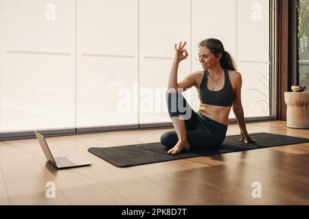 Fröhliche Seniorin, die während eines Online-Yoga-Kurses eine gute Geste macht. Glückliche Frau, die einem virtuellen Fitnesstutorial auf einem Laptop folgt. Reife Frau Prac Stockfoto