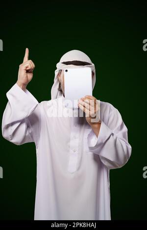 Moslemisches anonymes Konzept. Eine arabische Person hält ein digitales Tablet, das Gesicht bedeckt, den Zeigefinger nach oben zeigt und das Konzept der islamischen Zensur im Internet repräsentiert. Das Bild stellt Fragen der Privatsphäre, Sicherheit und Anonymität in der digitalen Welt dar. Hochwertiges Foto Stockfoto