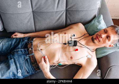 Ein Mann, der auf einem Sofa liegt, mit angeschlossenem Holter-Herzmonitor, Kardiologie Stockfoto