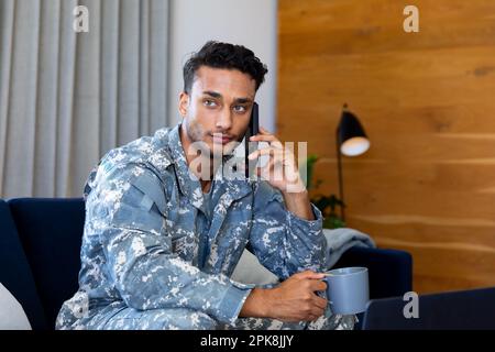 Rücksichtsvoller, birassistischer Soldat in Uniform, sitzt auf dem Sofa und spricht mit einem Smartphone im Wohnzimmer. Lifestyle, Militärdienst, Kommunikation und kostenlos Stockfoto