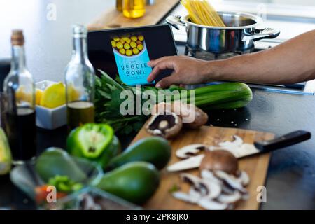 Nahaufnahme der Hände eines birassischen Mannes, der das Essen zubereitet, mit einem Tablet in der Küche. Lebensstil, Kochen, Kommunikation, Technologie und häusliches Leben, unaltere Stockfoto