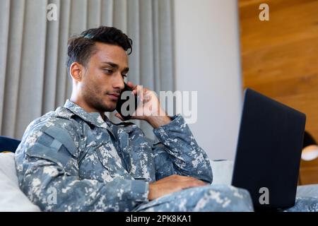 Birassischer Soldat trägt Uniform mit Laptop und spricht zu Hause über Smartphone. Lifestyle, Militärdienst, Kommunikation und Freizeit, unalt Stockfoto