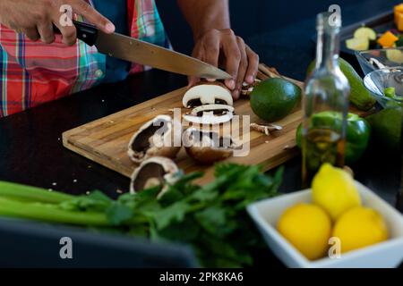 Nahaufnahme der Hände eines birassischen Mannes, der das Essen zubereitet, Pilze in der Küche hackt. Lebensstil, Kochen und häusliches Leben, unverändert. Stockfoto