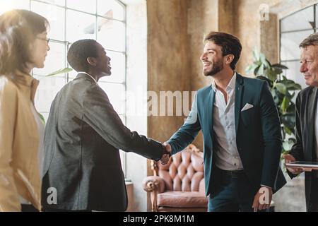 Junger weißer Mann und afroamerikanische Frau mit rasiertem Haar, lächelnd und händchenschüttelnd in einem modernen Büro, während Kollegen zuschauen. Tageslicht Stockfoto