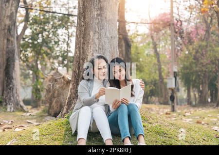 Eine reizende Enkelin und Großmutter, die im grünen Park unter dem Baum saßen, haben zusammen ein Buch gelesen Stockfoto