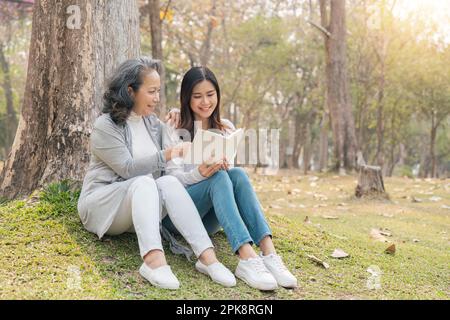 Eine reizende Enkelin und Großmutter, die im grünen Park unter dem Baum saßen, haben zusammen ein Buch gelesen Stockfoto