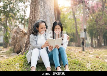 Eine reizende Enkelin und Großmutter, die im grünen Park unter dem Baum saßen, haben zusammen ein Buch gelesen Stockfoto