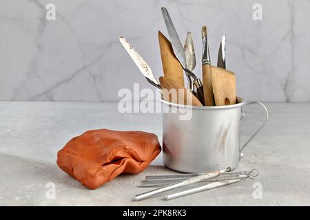 Eine Vielzahl von Werkzeugen zum Formen und Modellieren in einem Aluminiumbecher und einem Stück Tonplastik mit leichtem Werkstatthintergrund Stockfoto
