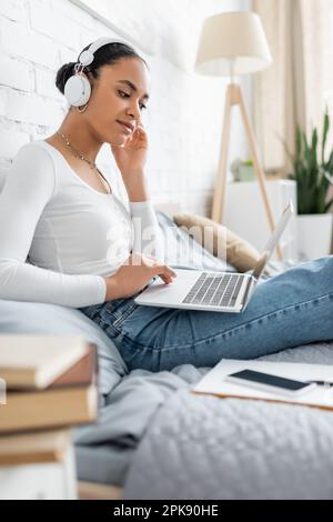 Junger afroamerikanischer Student mit kabellosen Kopfhörern, der ein Notebook benutzt, während er sich einen Vortrag im Internet ansieht, ein Stockbild Stockfoto