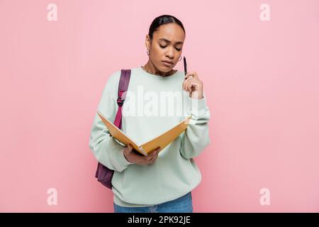 Frustrierte afroamerikanische Studentin, die Notizbuch und Stift in der Hand hielt, während sie mit einem Rucksack auf einem pinkfarbenen Stockbild stand Stockfoto