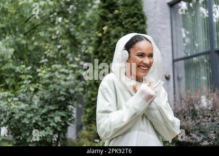 Eine überfreuliche afroamerikanische Frau in einem wasserdichten Regenmantel und kabellosen Kopfhörern, die unter Regen stehen, Stock Image Stockfoto