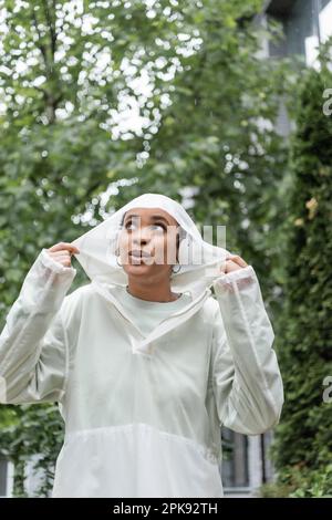 Überraschte afroamerikanische Frau in wasserdichter Regenjacke und kabellosen Kopfhörern, die unter Regen stehen, Stock Image Stockfoto