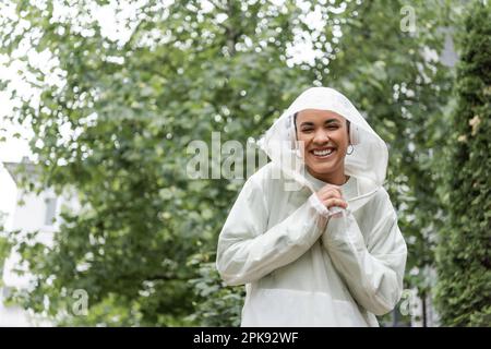 Fröhliche afroamerikanische Frau in wasserdichter Regenjacke und kabellosen Kopfhörern, die unter Regen stehen, Stock Image Stockfoto