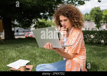 Pensiver Freiberufler mit lockigem Haar hält Laptop, während er im Green Park sitzt, Stockbild Stockfoto