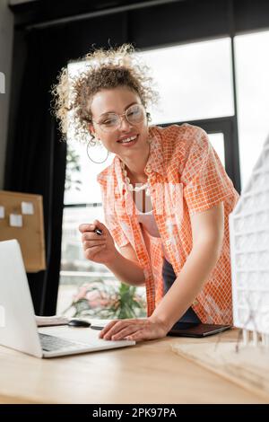 Glücklicher Architekturdesigner, der ein Notebook in der Nähe eines Grafiktabletts und ein Wohnmodell auf dem Schreibtisch verwendet, Stockbild Stockfoto