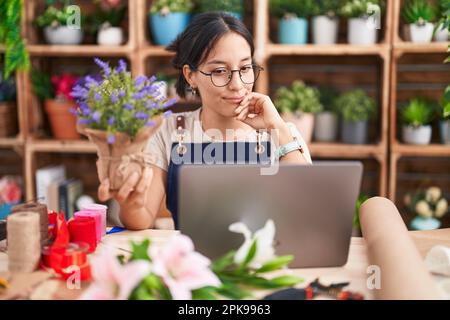 Junge hispanische Frau, die in einem Blumenladen arbeitet und Videoanrufe mit der Hand am Kinn macht und über Fragen nachdenklich ist. Lächelnder und aufmerksamer fa Stockfoto