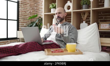 Ein junger Glatzkopf, der mit dem Laptop im Schlafzimmer frühstückt Stockfoto