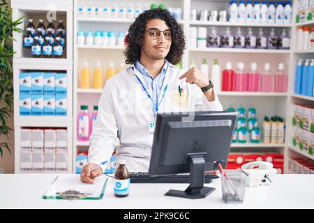 Hispanischer Mann mit lockigem Haar, der in der Apotheke arbeitet, selbstbewusst aussieht, mit einem Lächeln im Gesicht, sich mit stolzen und glücklichen Fingern zeigt. Stockfoto