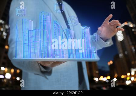 Ein Mann hält ein Tablet mit einem virtuellen Bild von Gebäuden und Ikonen vor verschwommenem Blick auf die nächtliche Stadtlandschaft, Nahaufnahme Stockfoto